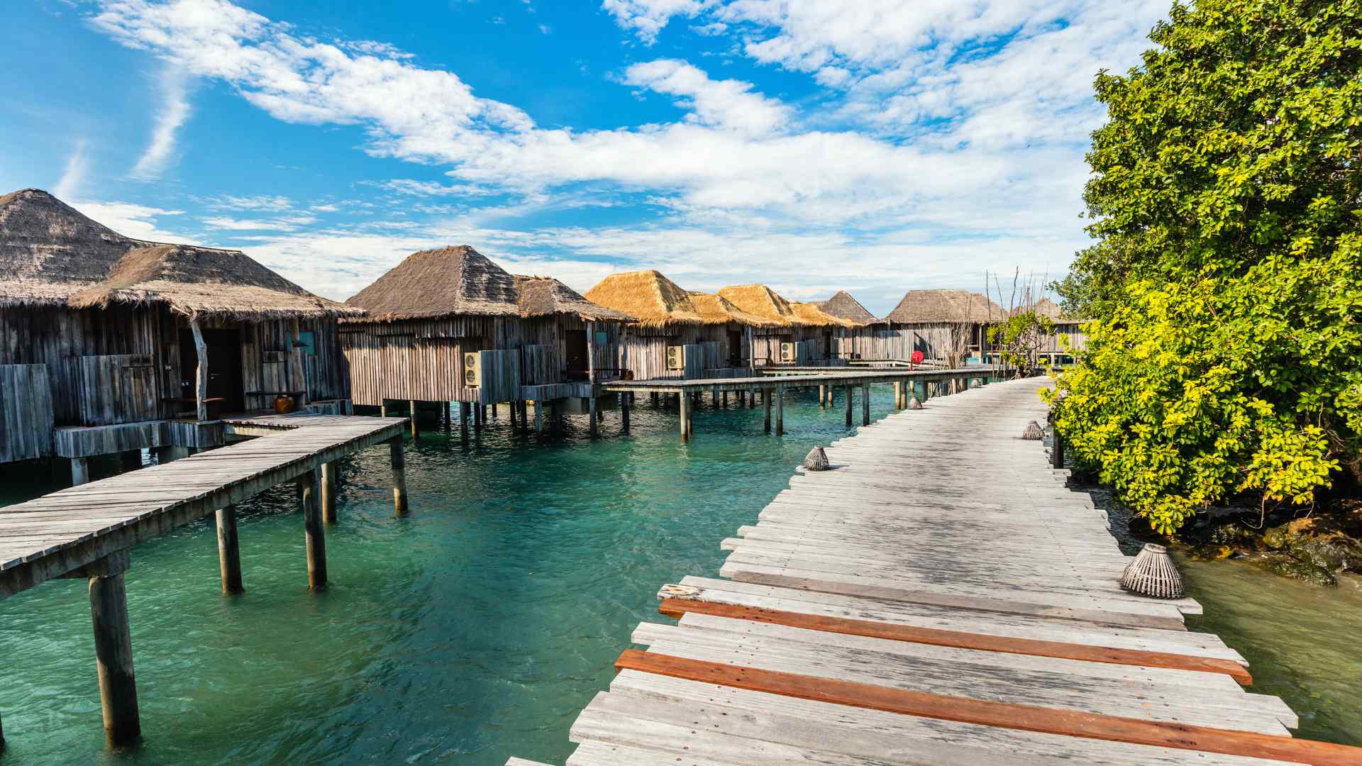 A thatched water villa with a wooden deck reaching out over shallow blue water